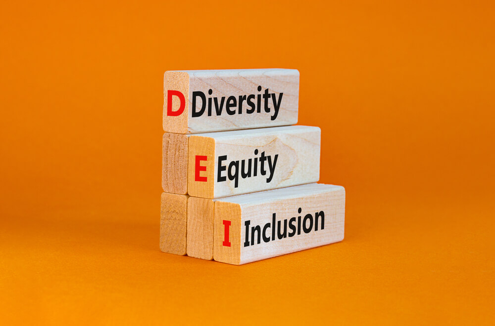 Stacked wooden blocks against an orange background display the words "Diversity," "Equity," and "Inclusion." The first letter of each word—D, E, I—is highlighted in red on the side of each block, forming the acronym "DEI," symbolizing the integration of AI into fostering inclusive environments.